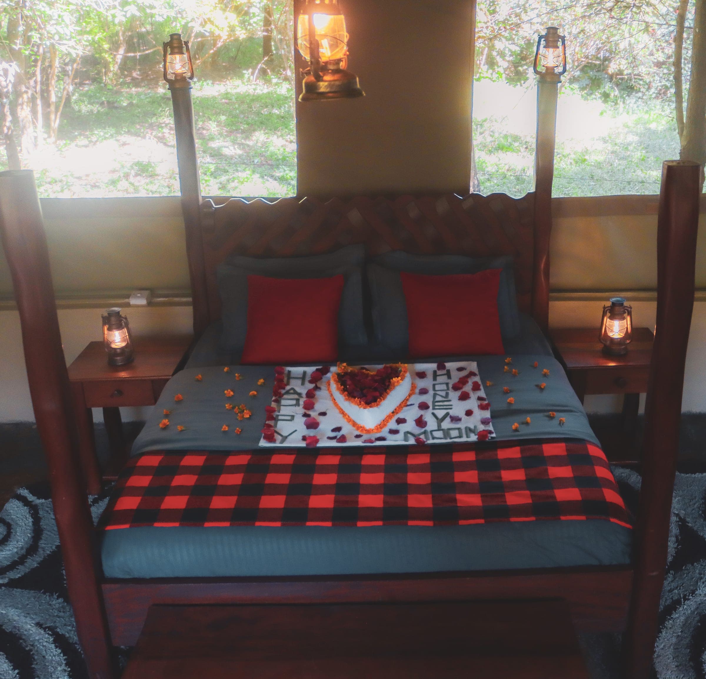 Four-poster bed set for honeymooners with a petal heart, red check blanket and lit lanterns, bush through the windows